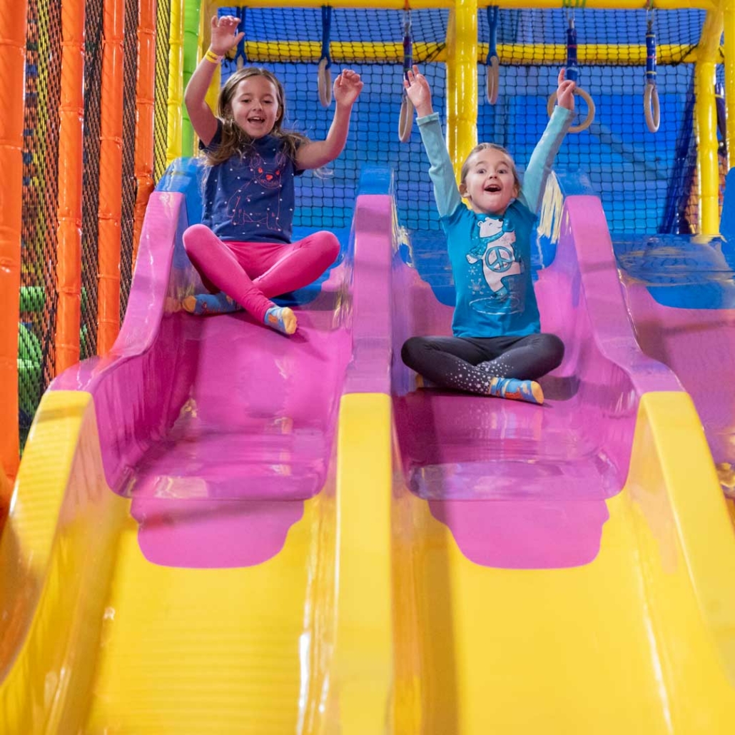 two girls sliding down a slide at an indoor playground